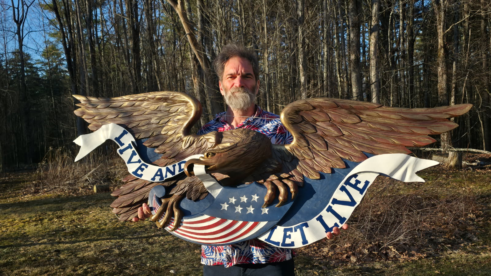 Craftsman holding a hand-carved gilded eagle 'Live and Let Live' sign in the woods